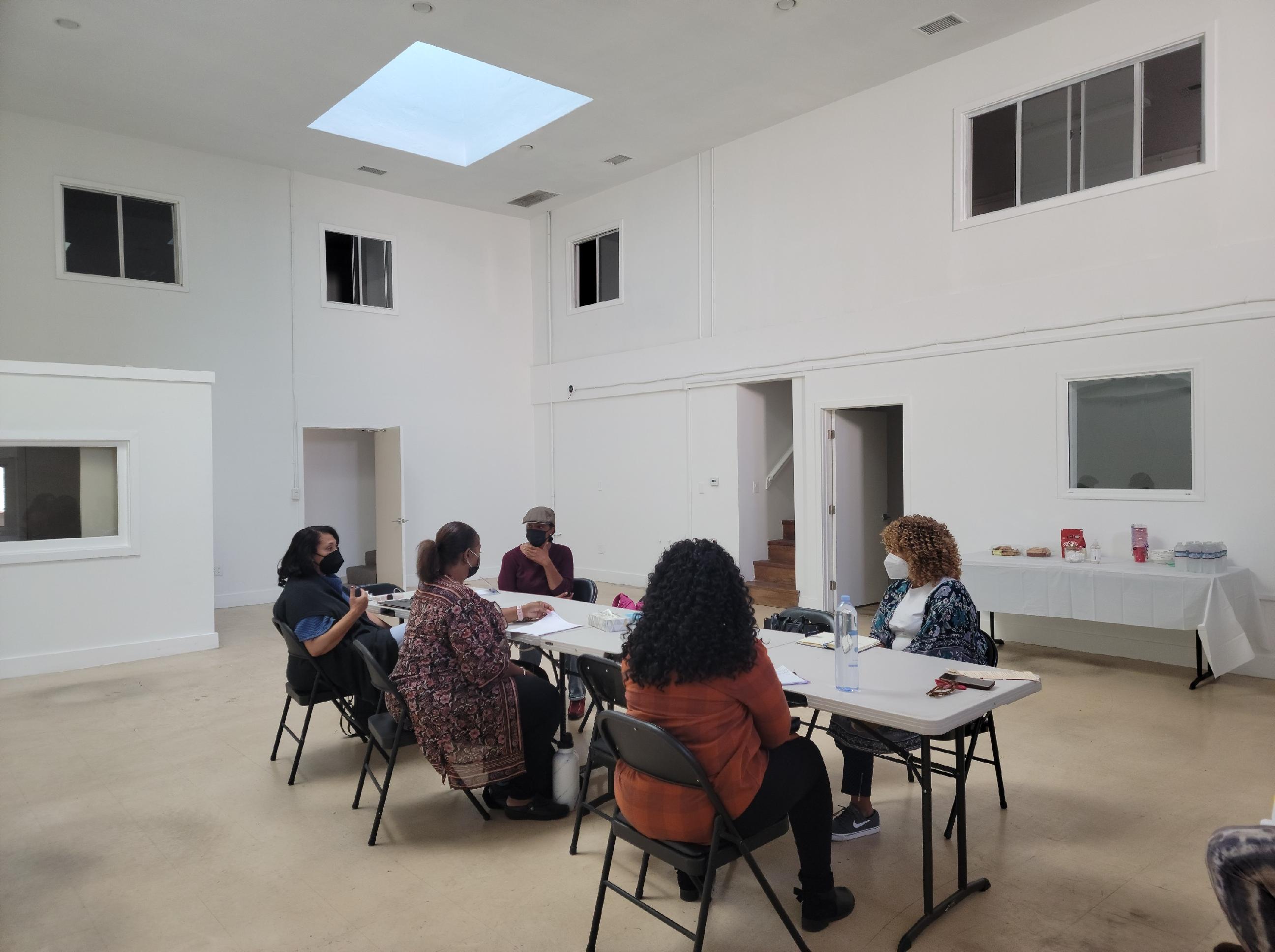 Five women in masks sit on around two folding tables in a large white room.