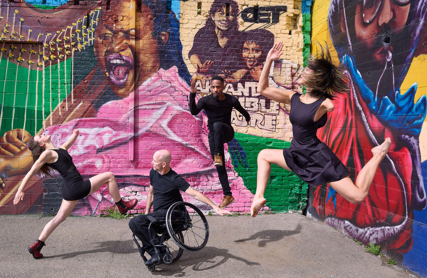 Man in wheelchair and three others wearing all black dancing in front of a colorful painted mural outside.