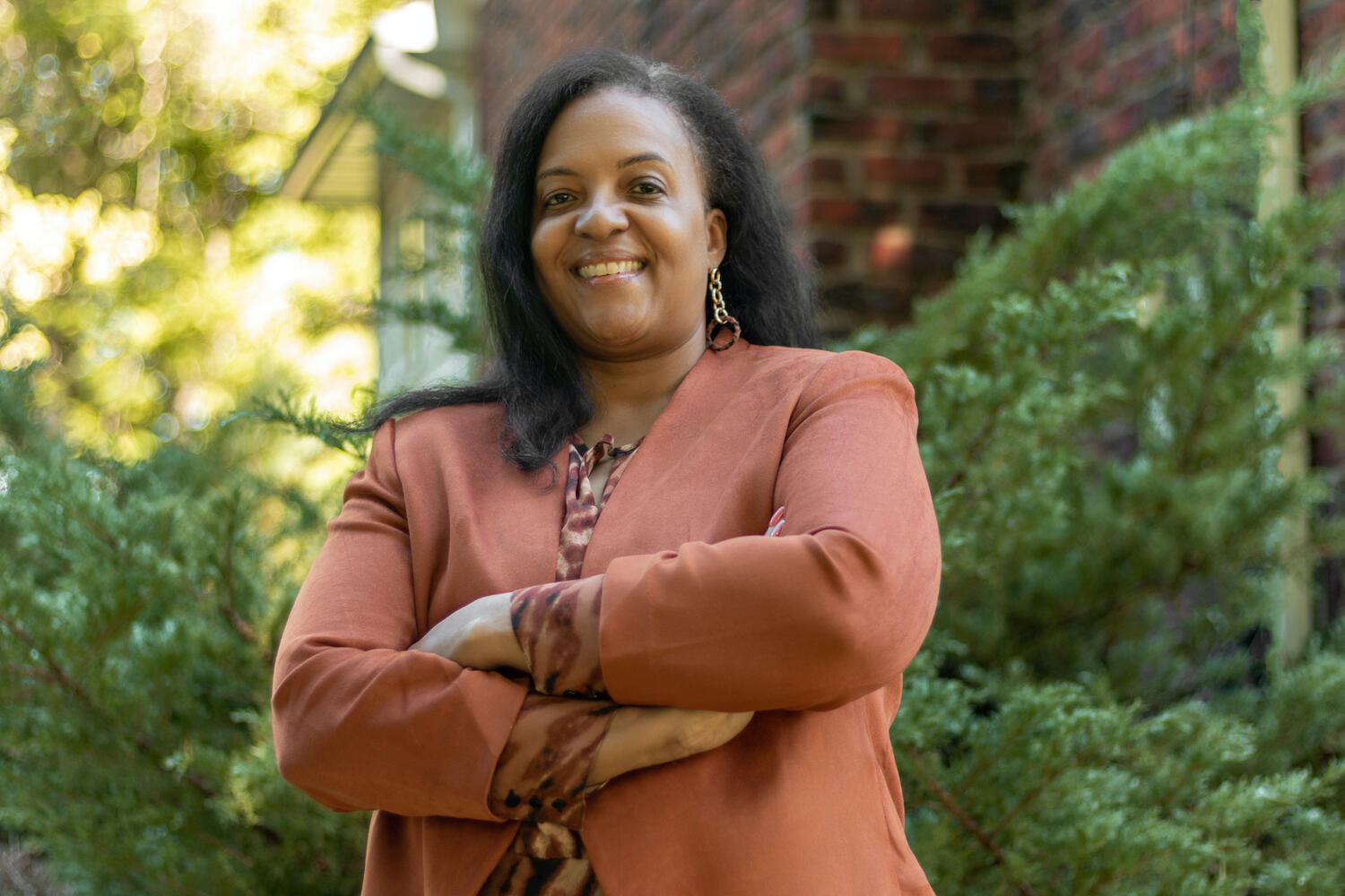 Headshot of Aisha standing outside in front of green bushes wearing a terra cotta colored blazer