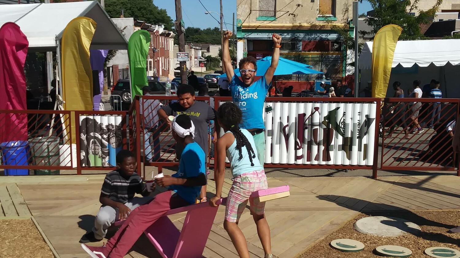 Three children sit on a seesaw, two are holding cups with food. Another child is approaching them and an adult with glasses is cheering against the backdrop of colorful siding that encloses a playground.