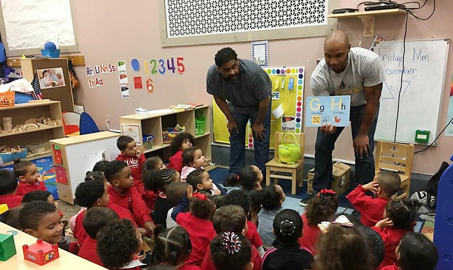 Two men of color, co-founders Akeiff Staples and Brent Johnstone, standing with their hands on their knees, showing an alphabet book, in front of a class of children of color sitting on the floor.