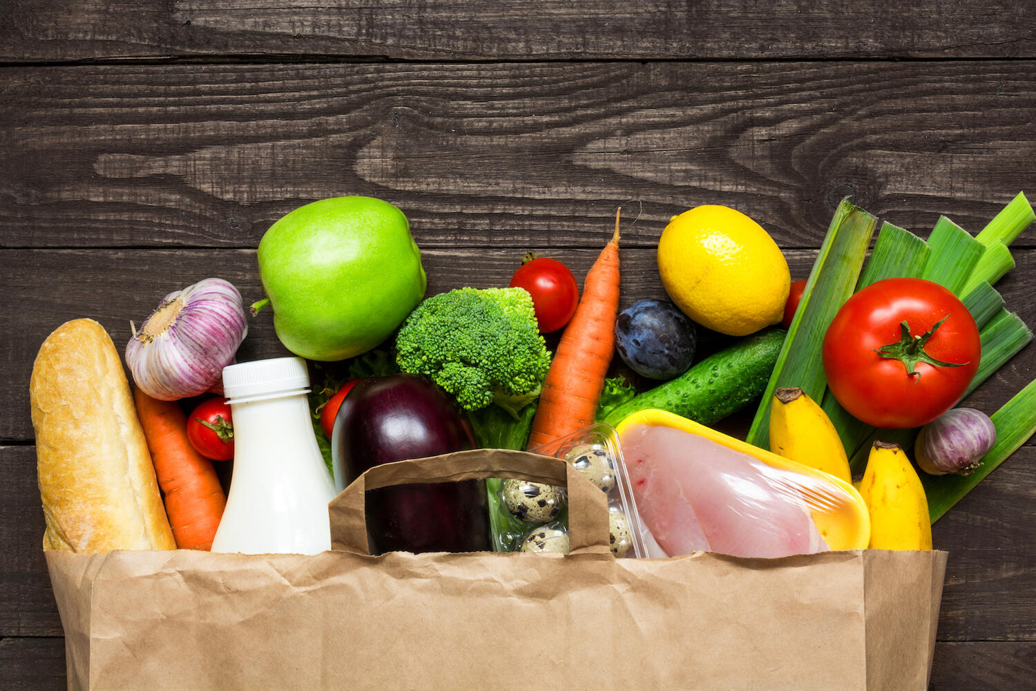 A paper bag laying flat, on a wooden surface with food spilling out. Items include, from left to right, a loaf of bread, carrot, garlic bulb, a white bottle, green apple, eggplant, broccoli, tomato, a container of quail eggs, a package of chicken, lemon, leeks, bananas