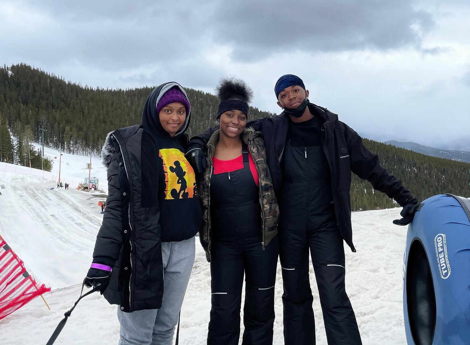 Three young people dressed in winter coats pose with an inner tube on a snowy mountain