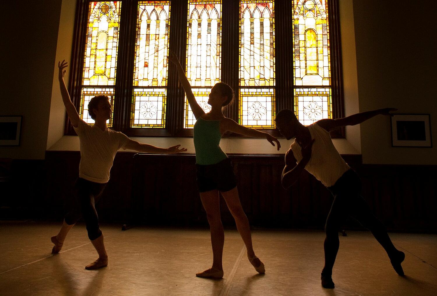 Three ballet dancers stretching. The dancers are backlit, so they appear in silhouette 