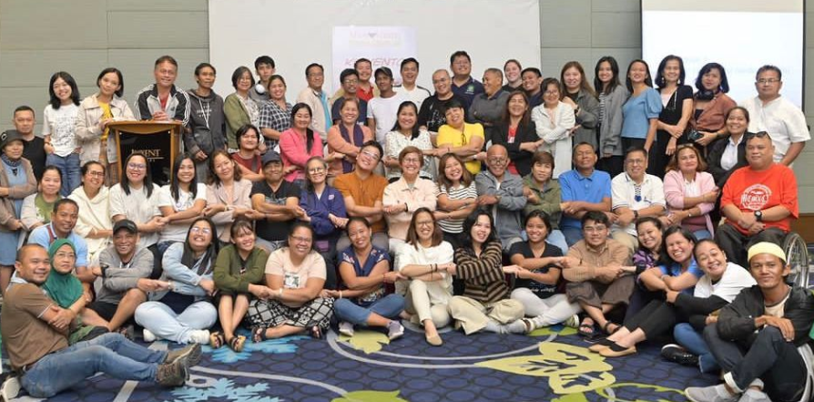 A large group of participants poses for a photograph in a hotel conference room with a colorful blue and yellow carpet.