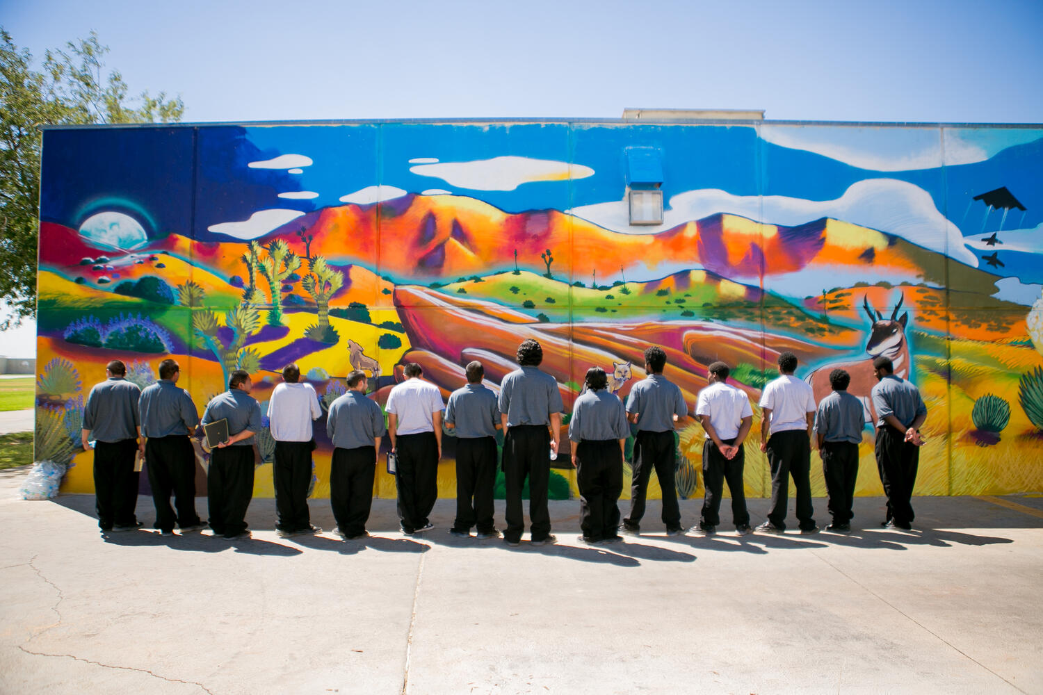 A group of young men stand facing a colorful mural with a blue sky, clouds, rolling hills, and red mountains. They are wearing white and grey tops and black pants.