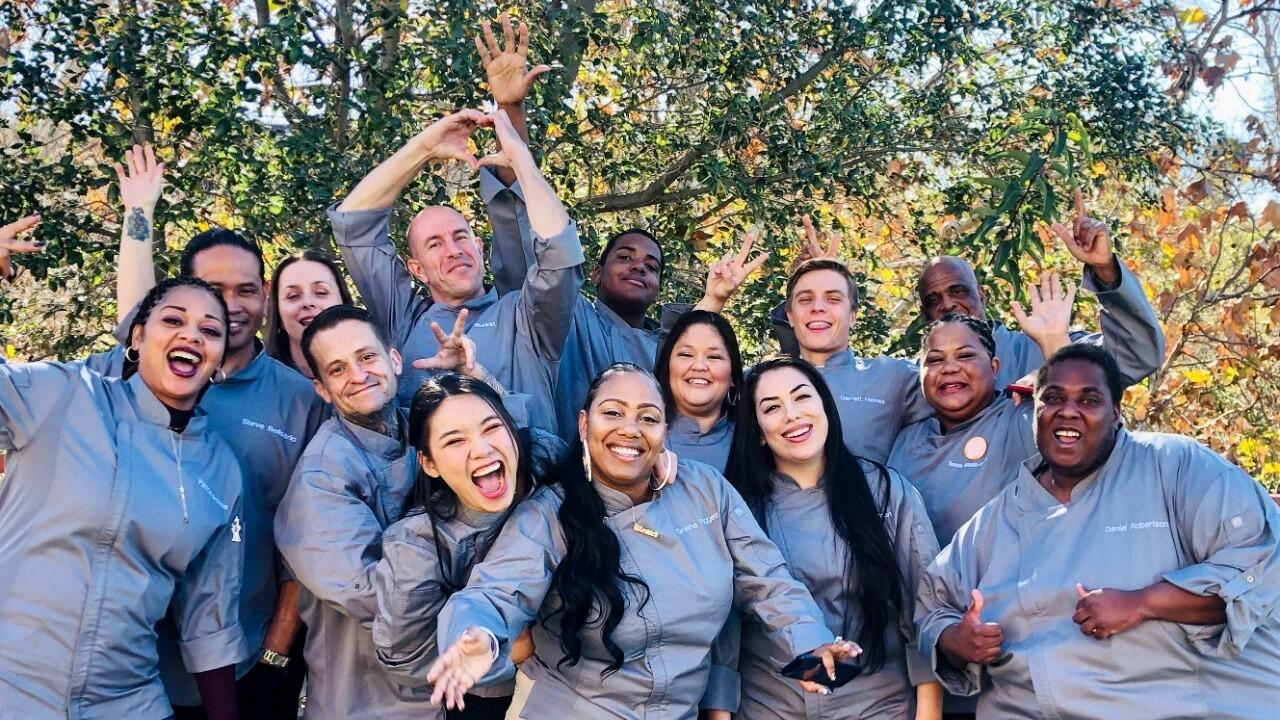 Group of people from diverse racial backgrounds standing outside in front of a tree, all wearing gray long sleeved shirts, smiling and waving at the camera.