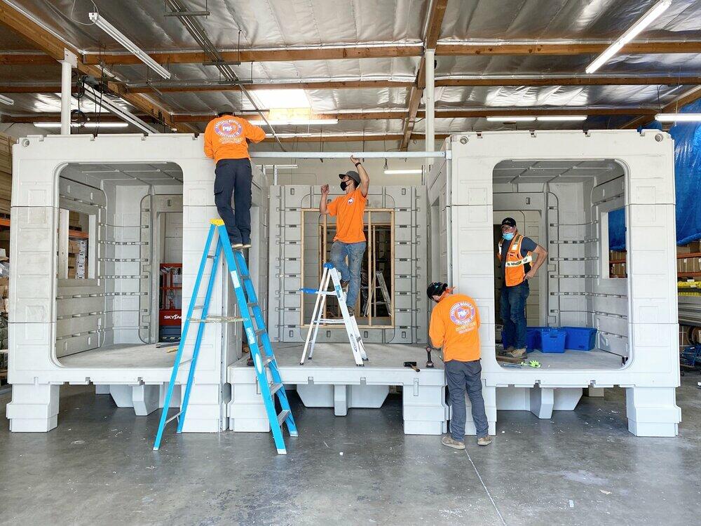 Four people in orange shirts stand inside and next to three large, plastic boxes