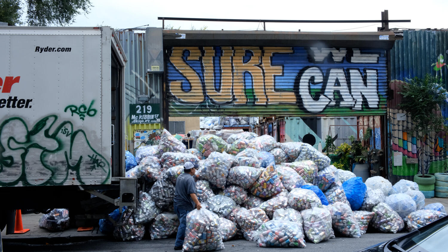 Director of Operations Rene Del Carmen at the entrance to Sure We Can on McKibbin St in Brooklyn New York in front of huge pile of plastic bags containing aluminum cans next to a large truck