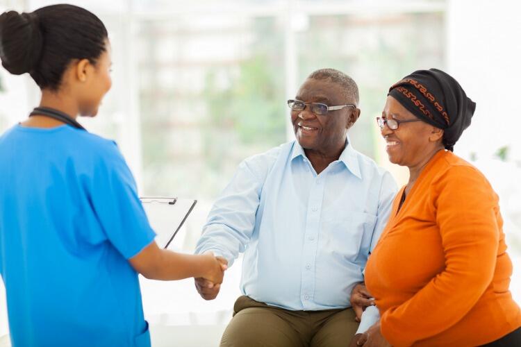 An older couple of color sitting on the right, shaking hands with a woman of color in blue hospital scrubs.