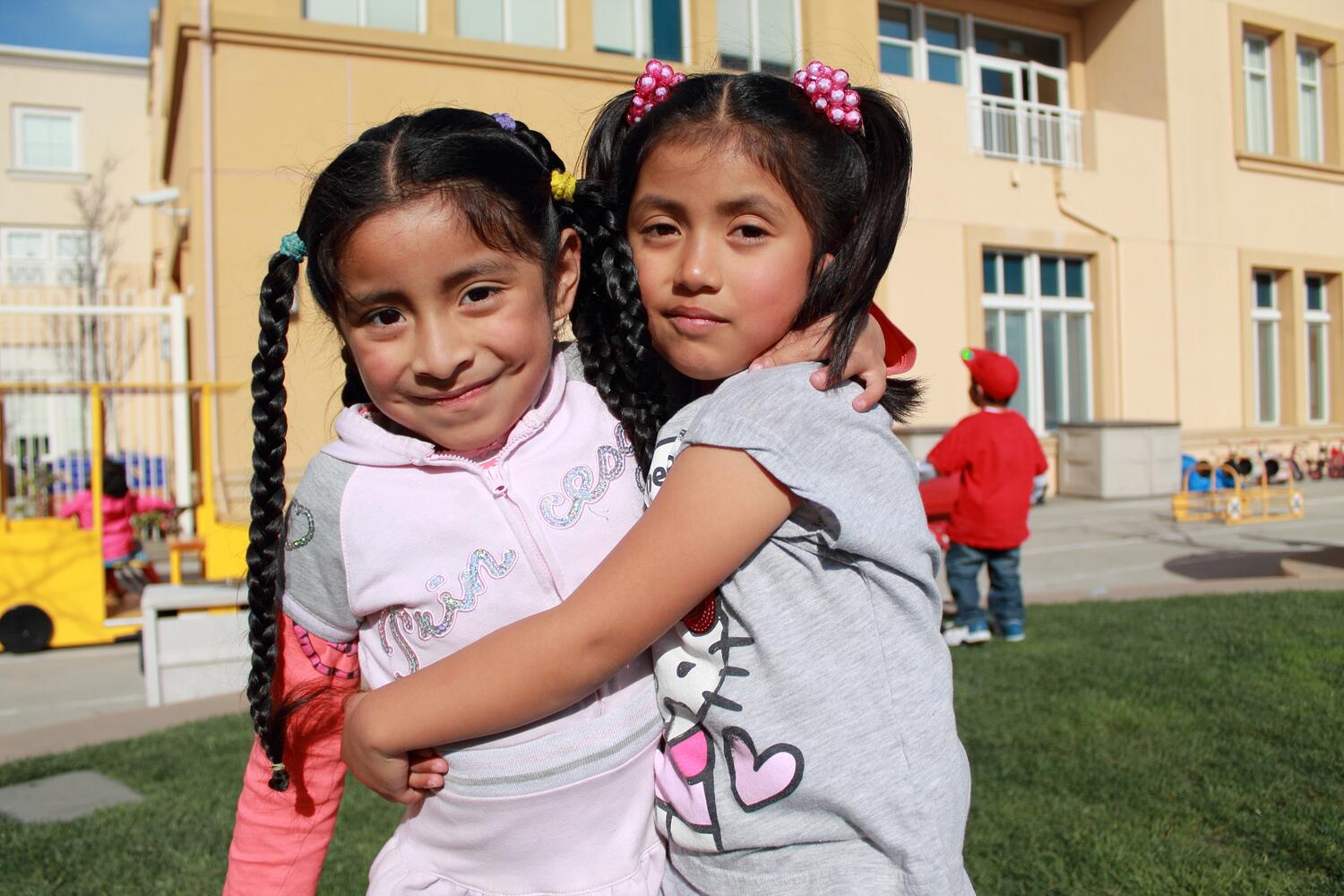 A close up of two young girls of color, hugging each other outside on a sunny day, facing forward. The girl on the left has pigtails and is wearing a pink jacket. The girl on the right has ponytails and is wearing a gray shirt. 