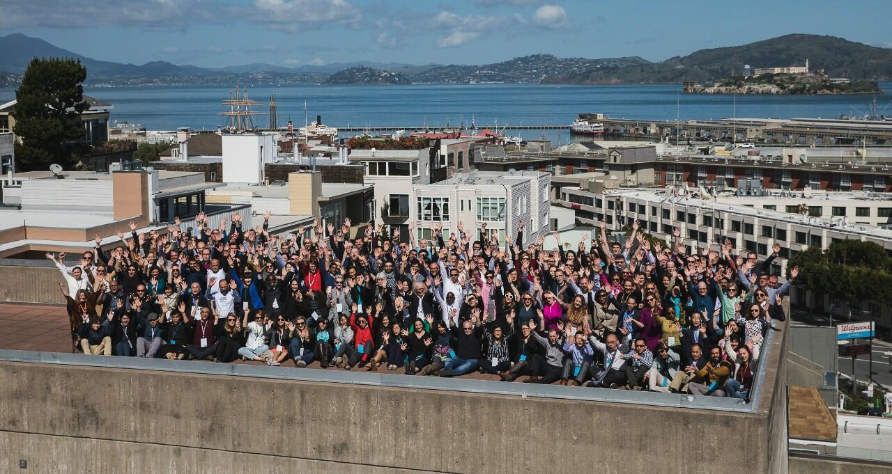 Large group of people sitting and standing on top of a building with ocean and mountains in the background.