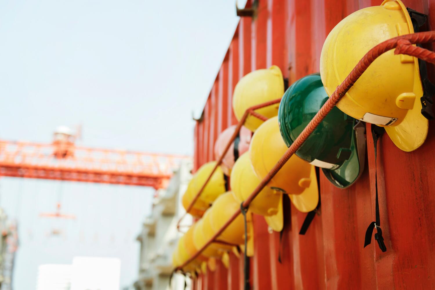 Image features a row of hard hats lined up on a container with a crain off in the distance. Photo by rawpixel.com from Pexels