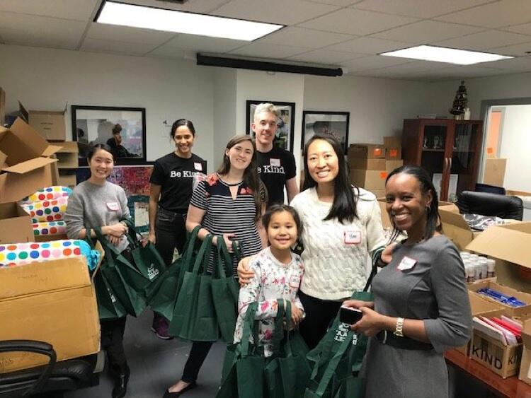 Volunteers at The Ali Forney Center. There are seven people holding green totebags, and they are surrounded by boxes of supplies