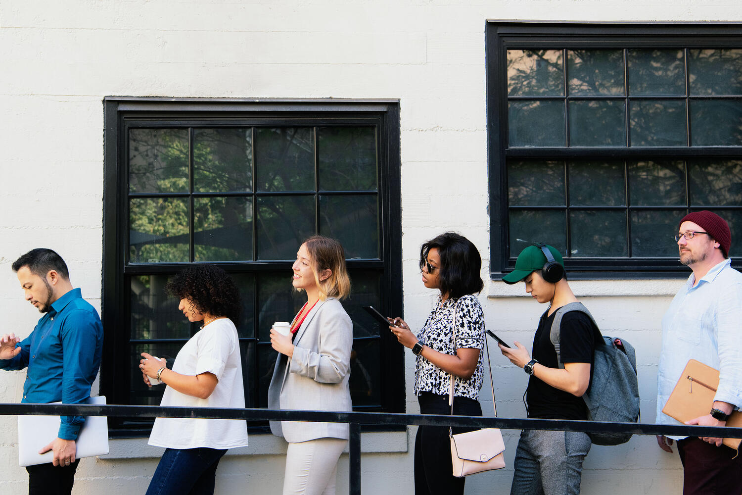 People standing in line next in front of a white building with black windows; one person is checking their watch, many are looking at their phones.