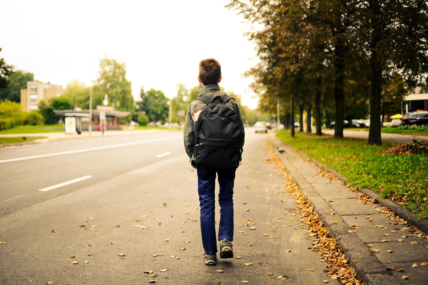 The back of a young boy walking on the side of a street. He's wearing a backpack, a jacket and dark blue jeans. 