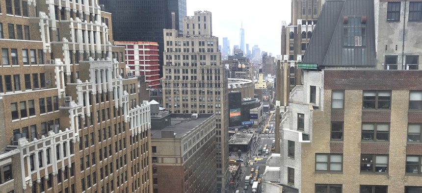 View of NYC skyline from the top of a building looking out over a busy street and cascading rooftops.