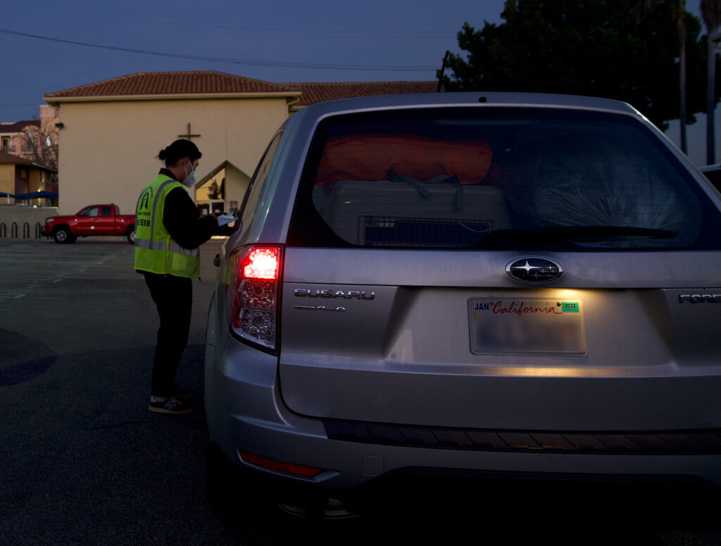 A person in a yellow vest looking at a clipboard, next to a parked car.