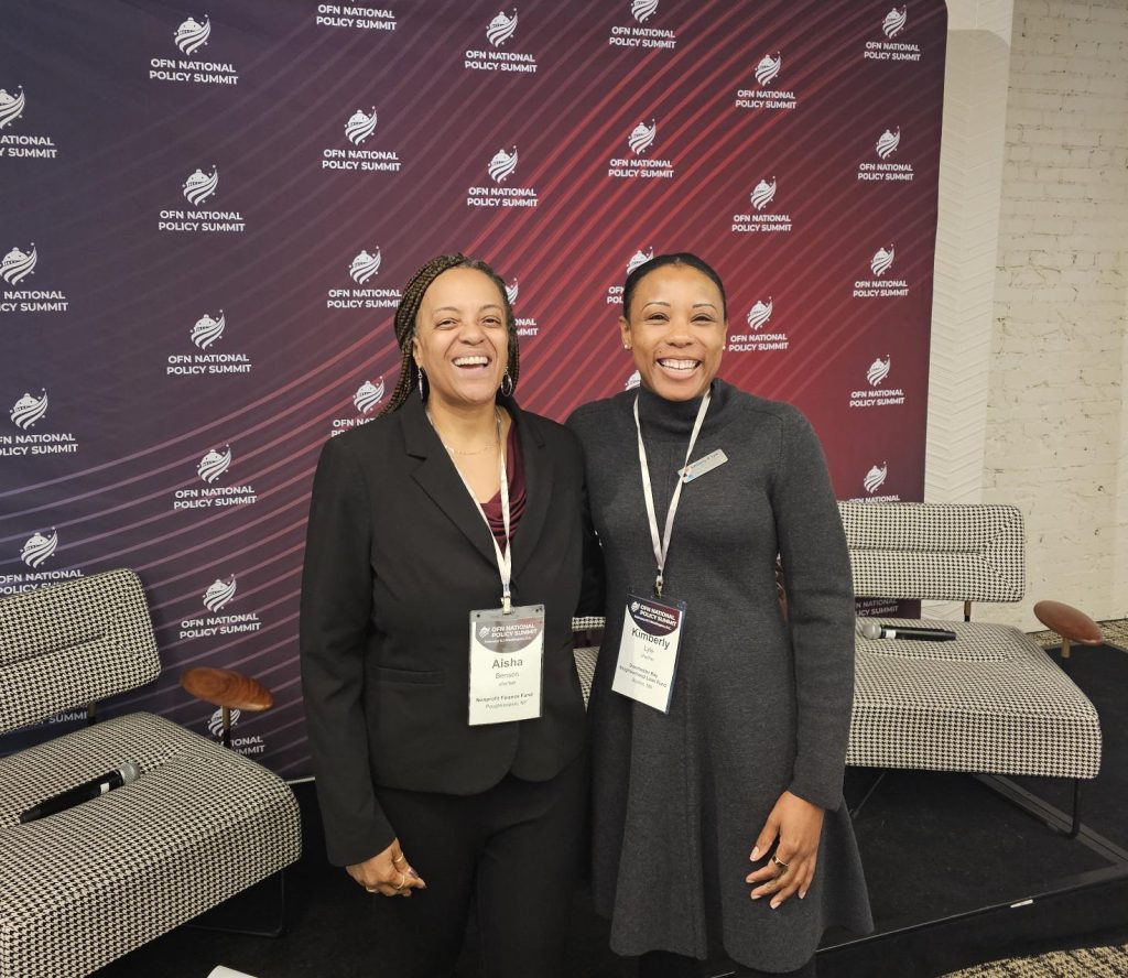 Aisha Benson and Kimberly Lyle smiling at the camera in front of a red and purple ombre sign that says OFN National Policy Summit.