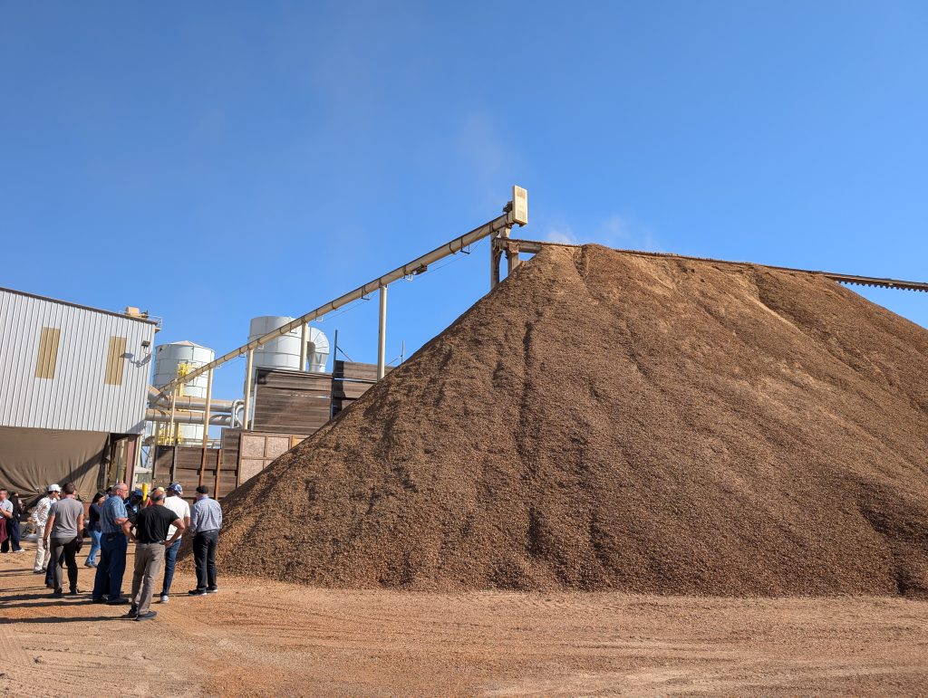 Large pile of almond shells in front of a processing facility with a group of people standing to the left.