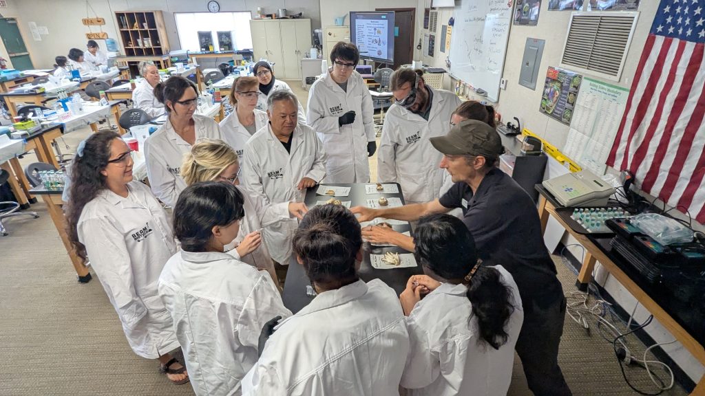 A group of people wearing white lab coats gather around a table where an instructor is explaining different materials.