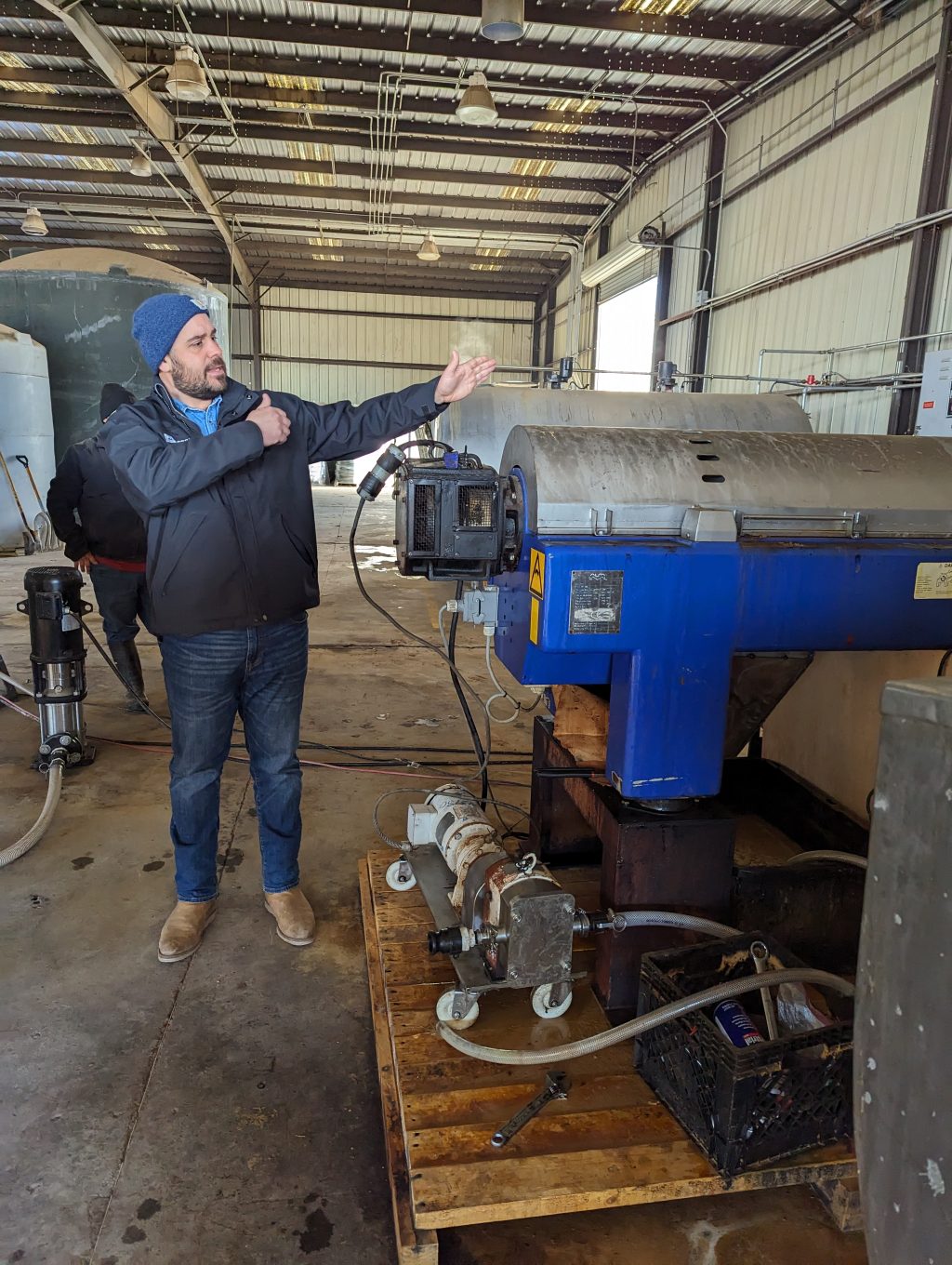 A person wearing dark blue jacket and jeans has arms raised to point out features of a piece of machinery to his left.