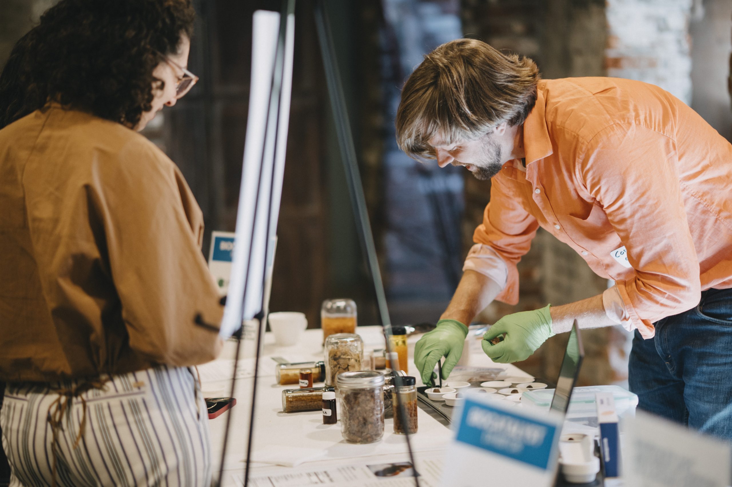 Person wearing an orange shirt and green gloves working with seeds and organic materials on a table; another person is looking at his work.