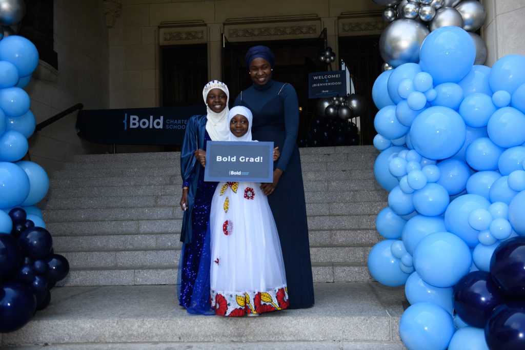 A Bold graduate stands proudly with their family at 5th grade graduation. A blue balloon arch surrounds them.