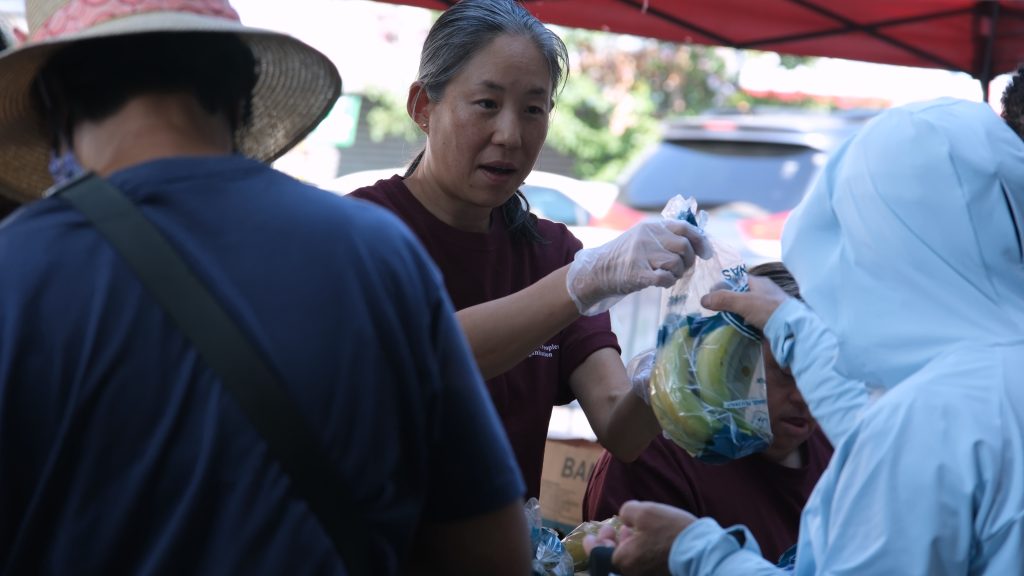 A woman hands a clear bag of bananas to a person whose face is obscured.