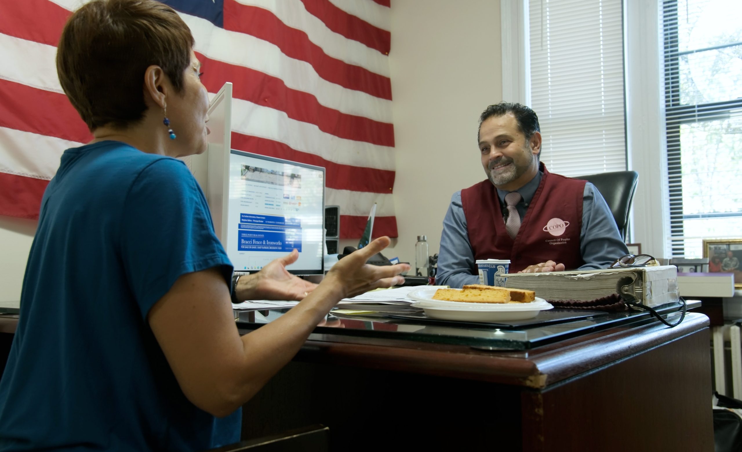 Ines and Moe sit across a desk in an office, engaged in a focused conversation with a computer and an American flag in the background.