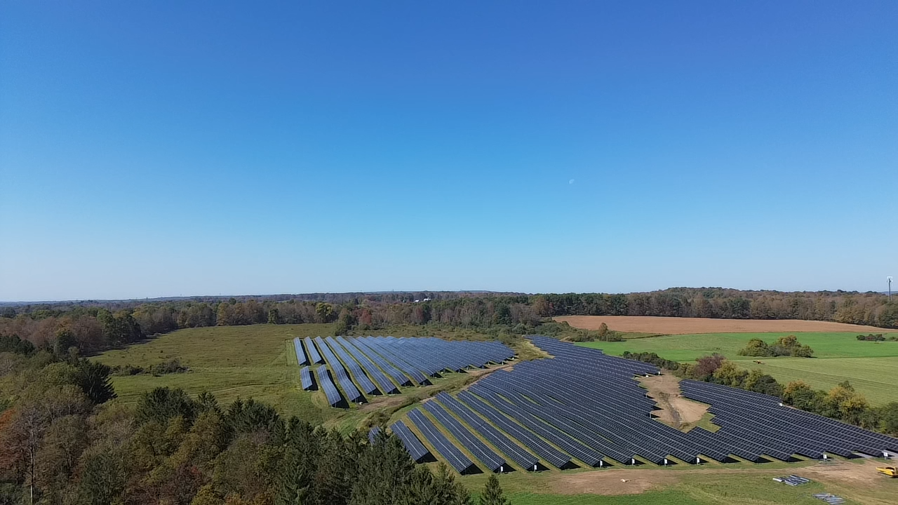 Aerial view of a solar farm with rows of solar panels in a green field, surrounded by trees under a clear blue sky. The scene conveys a sense of sustainability and innovation.