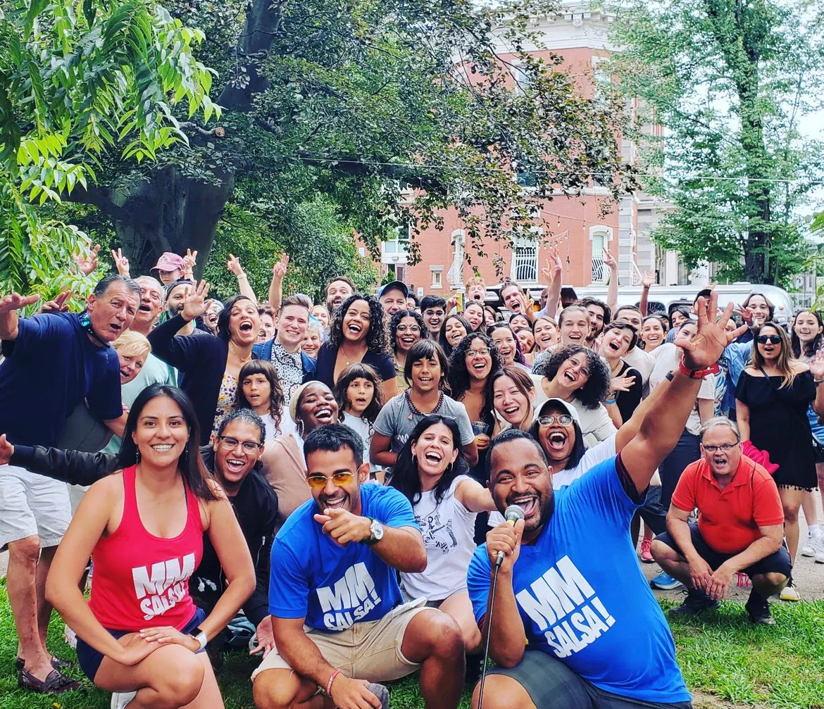 A large, diverse group of people smiles and cheers for a group photo in an outdoor park. In the foreground, people wearing "MM SALSA!" t-shirts kneel and smile, with one man holding a microphone.