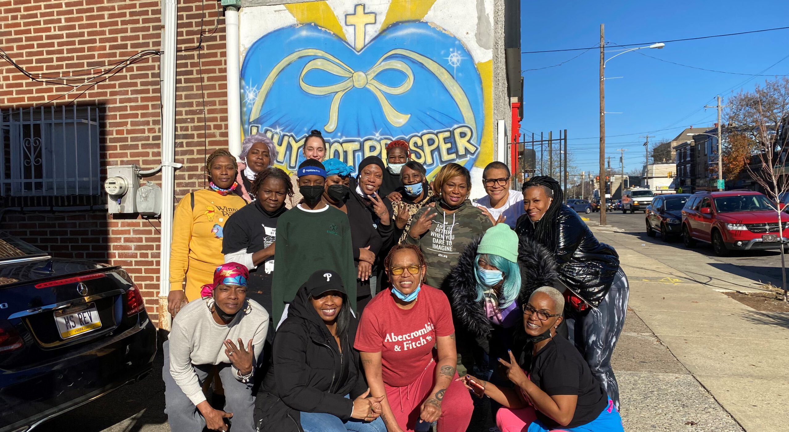 A large group of women smile and pose for a group photo on a sunny city sidewalk. They are gathered in front of a brick building with a large mural of a blue heart with a yellow bow.