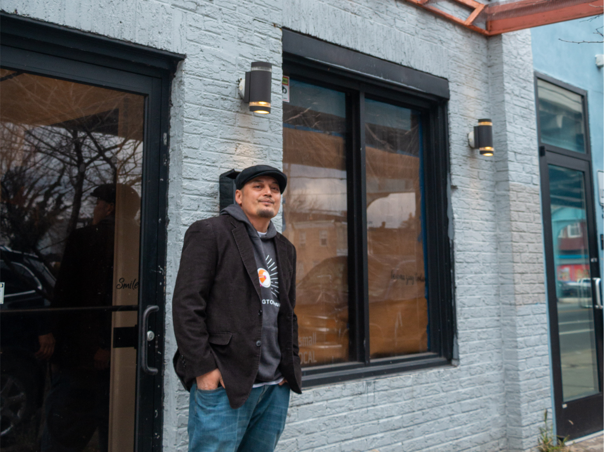 Man stands in front of a light blue brick building with a metal awning and the number 3312. Dressed casually, he appears relaxed and content.
