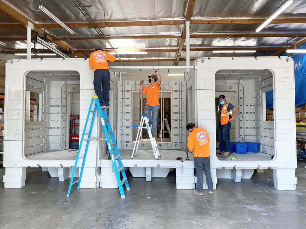 Four construction workers in orange shirts use ladders and tools to assemble a large, white, prefabricated structure inside a warehouse.