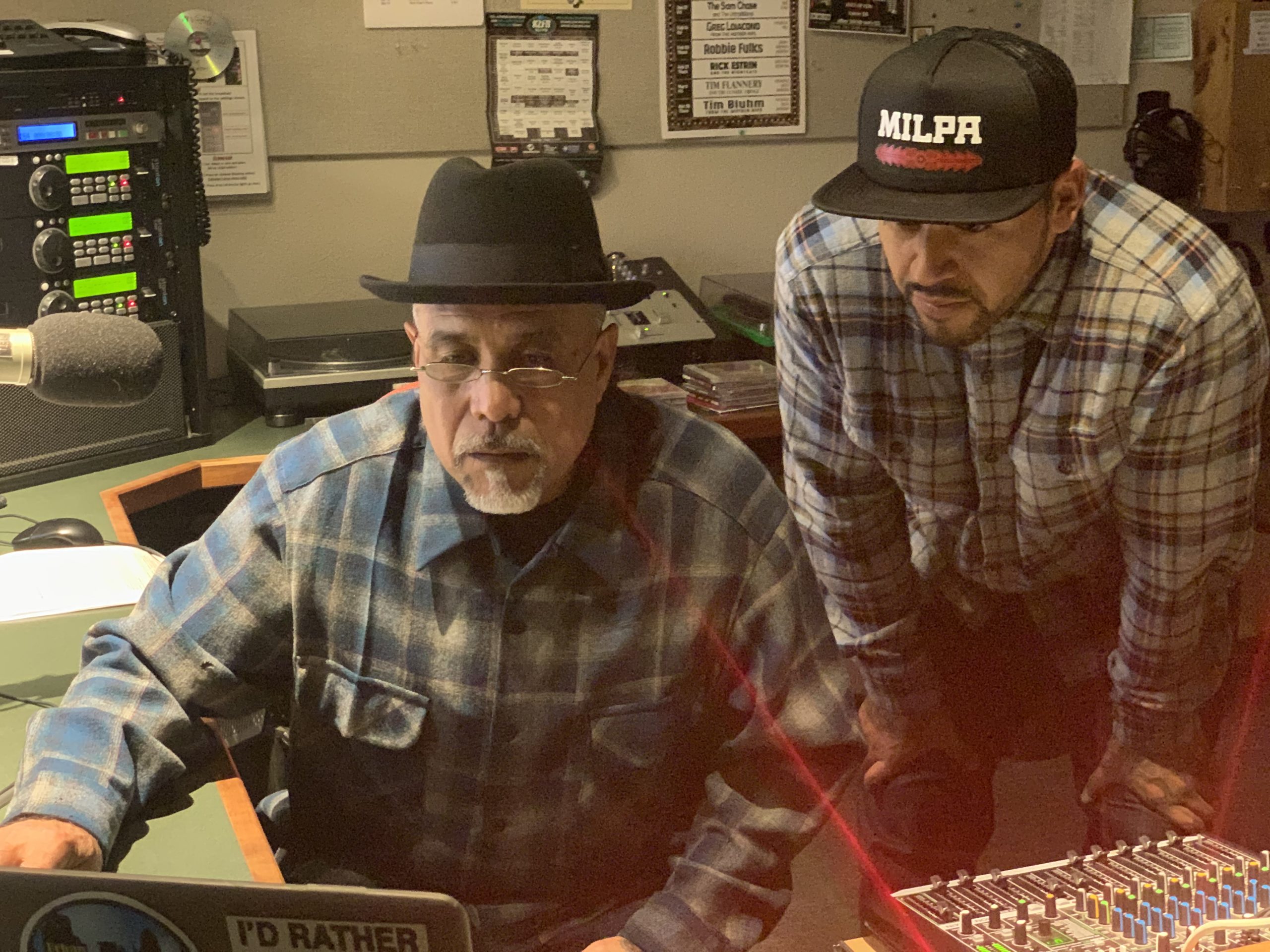 Two men looking at a computer screen in a room with a sound mixer board, a microphone, and other audio equipment.