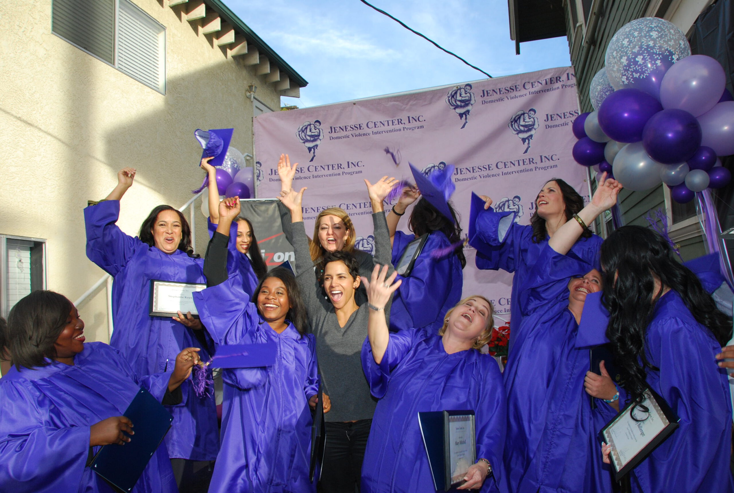 A group of women in purple graduation gowns joyfully celebrate outdoors, holding certificates and throwing their caps in the air. They stand in front of a pink "Jenesse Center, Inc." banner and purple balloons.