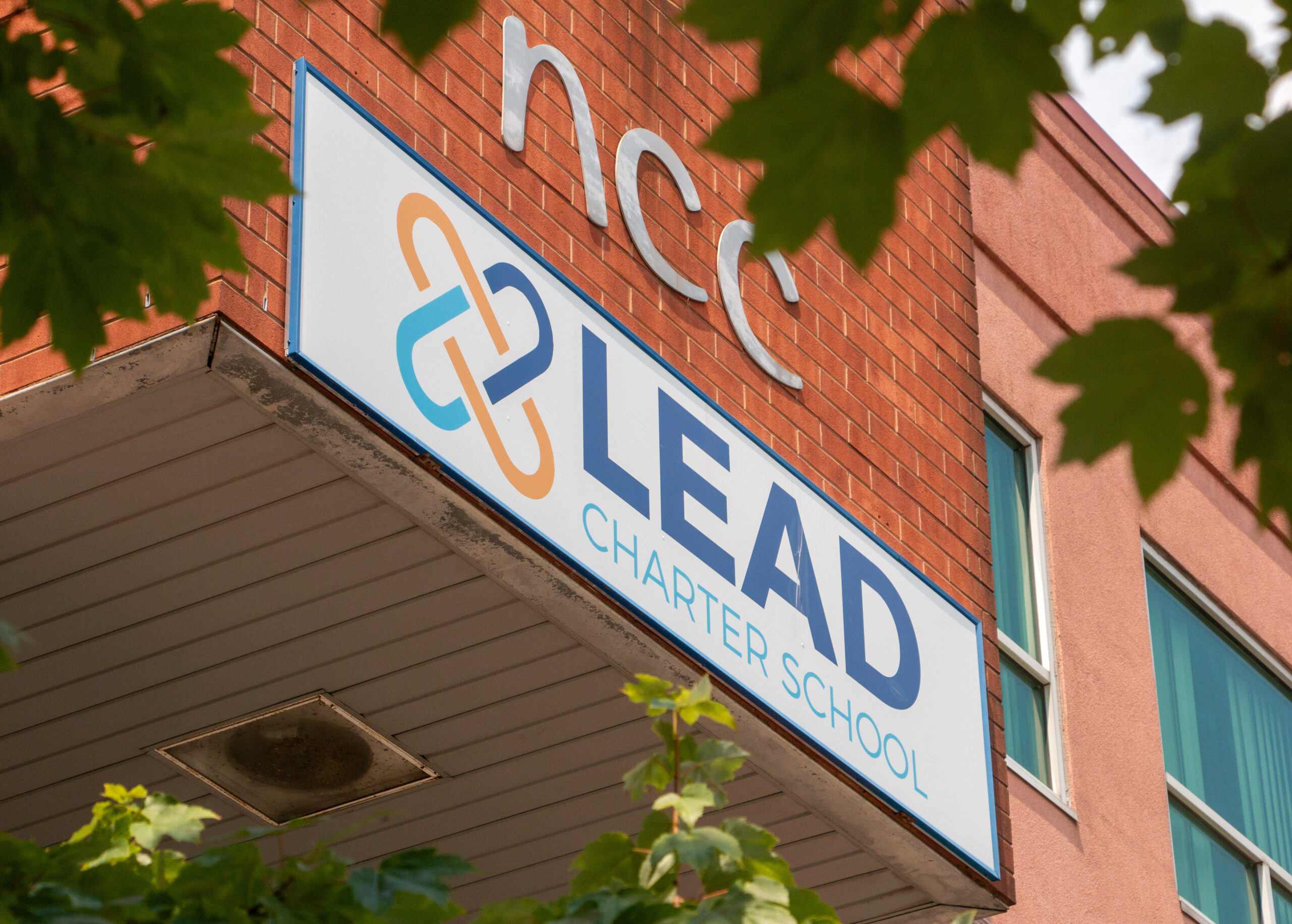 Sign for Lead Charter School on a brick building, partially obscured by green leaves. The sign features a logo and the text is clear and bold.