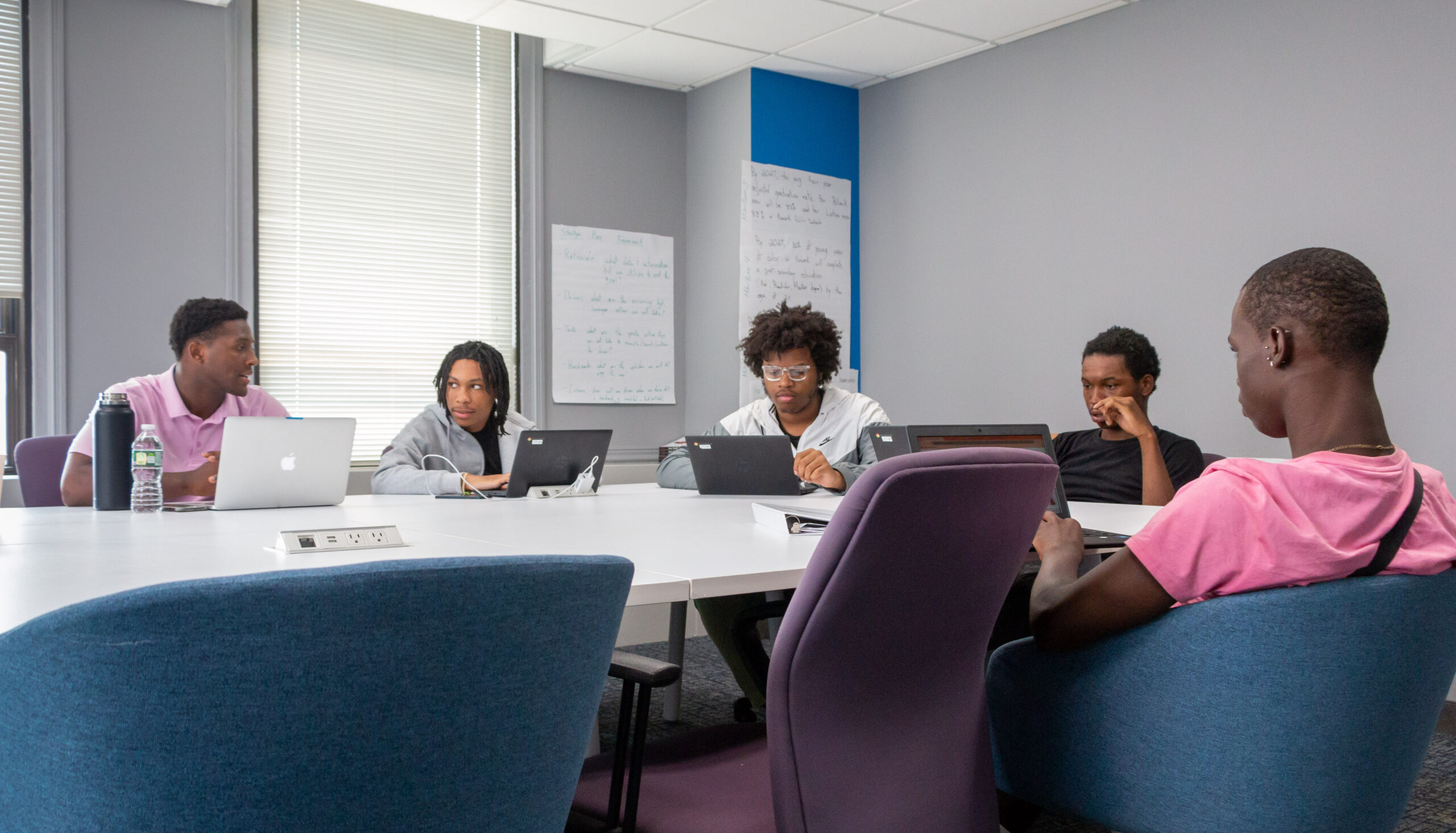 Five people sit around a conference table with laptops, engaged in discussion. The room has a focused, collaborative atmosphere. Whiteboards are in the background.