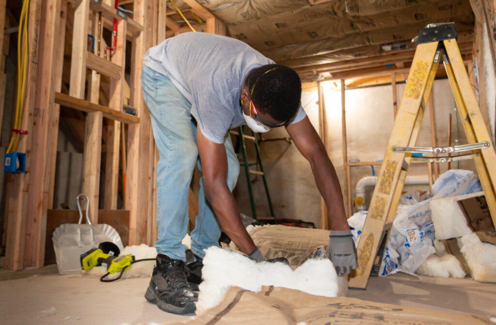 A person in protective goggles and mask installs insulation in a partially constructed room, conveying focus and productivity. A ladder and tools are nearby.