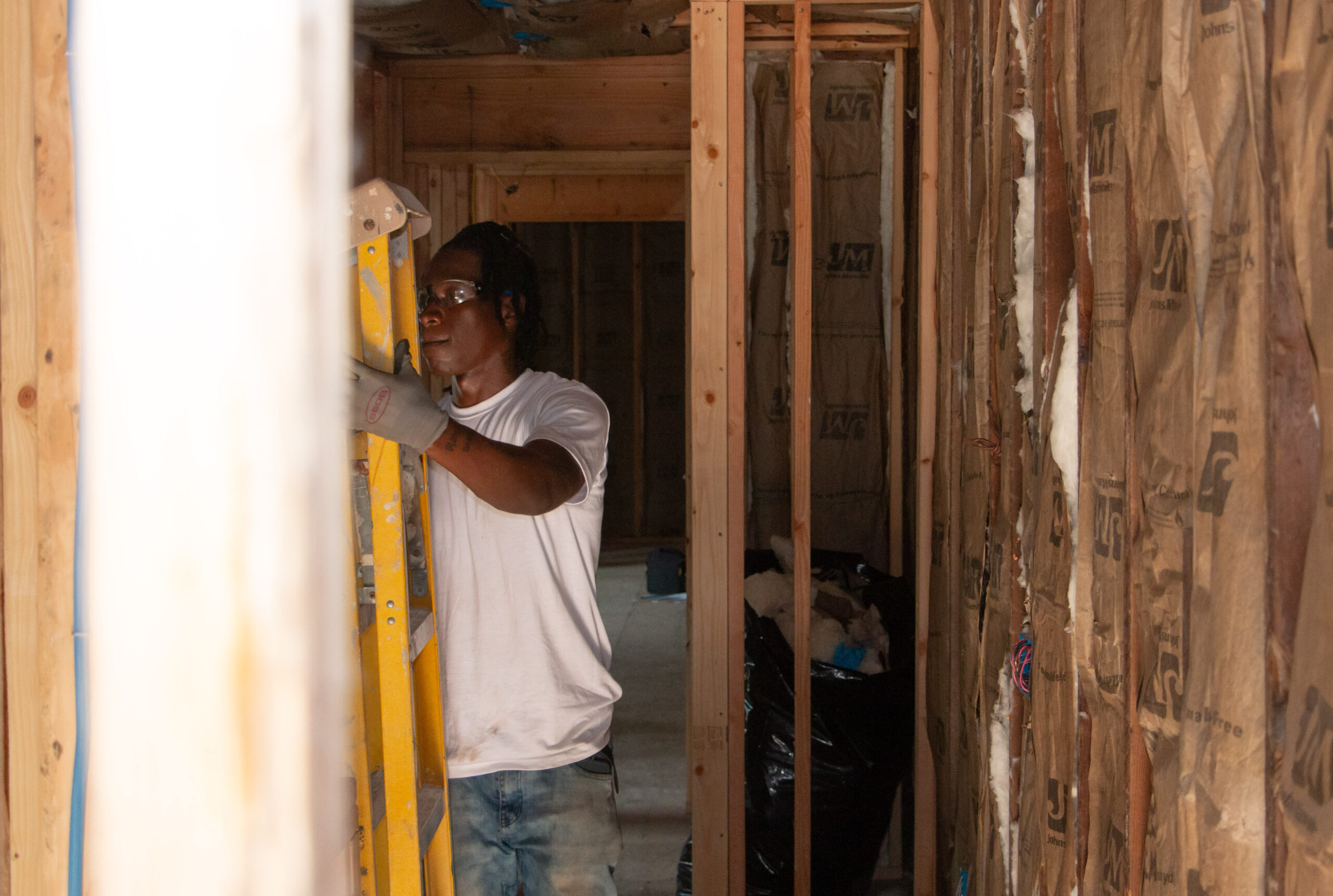A person wearing a white shirt and safety glasses stands on a yellow ladder in an unfinished room with exposed wooden beams and insulation. The scene conveys concentration and construction work in progress.