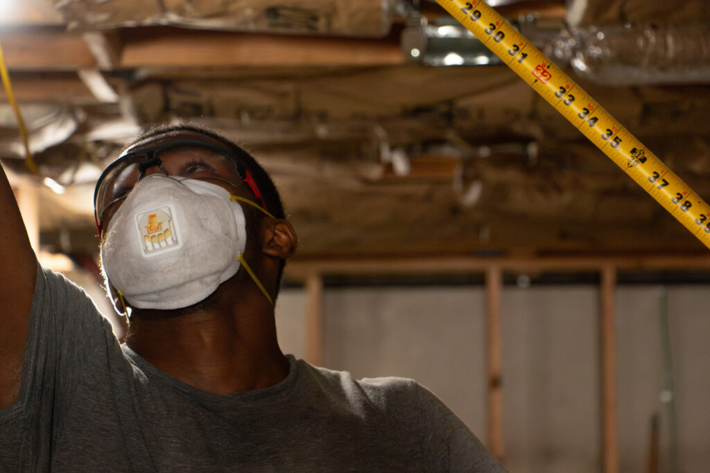 Man in a construction setting wears a mask and goggles, holding a tape measure against an unfinished ceiling, focused and attentive to detail.