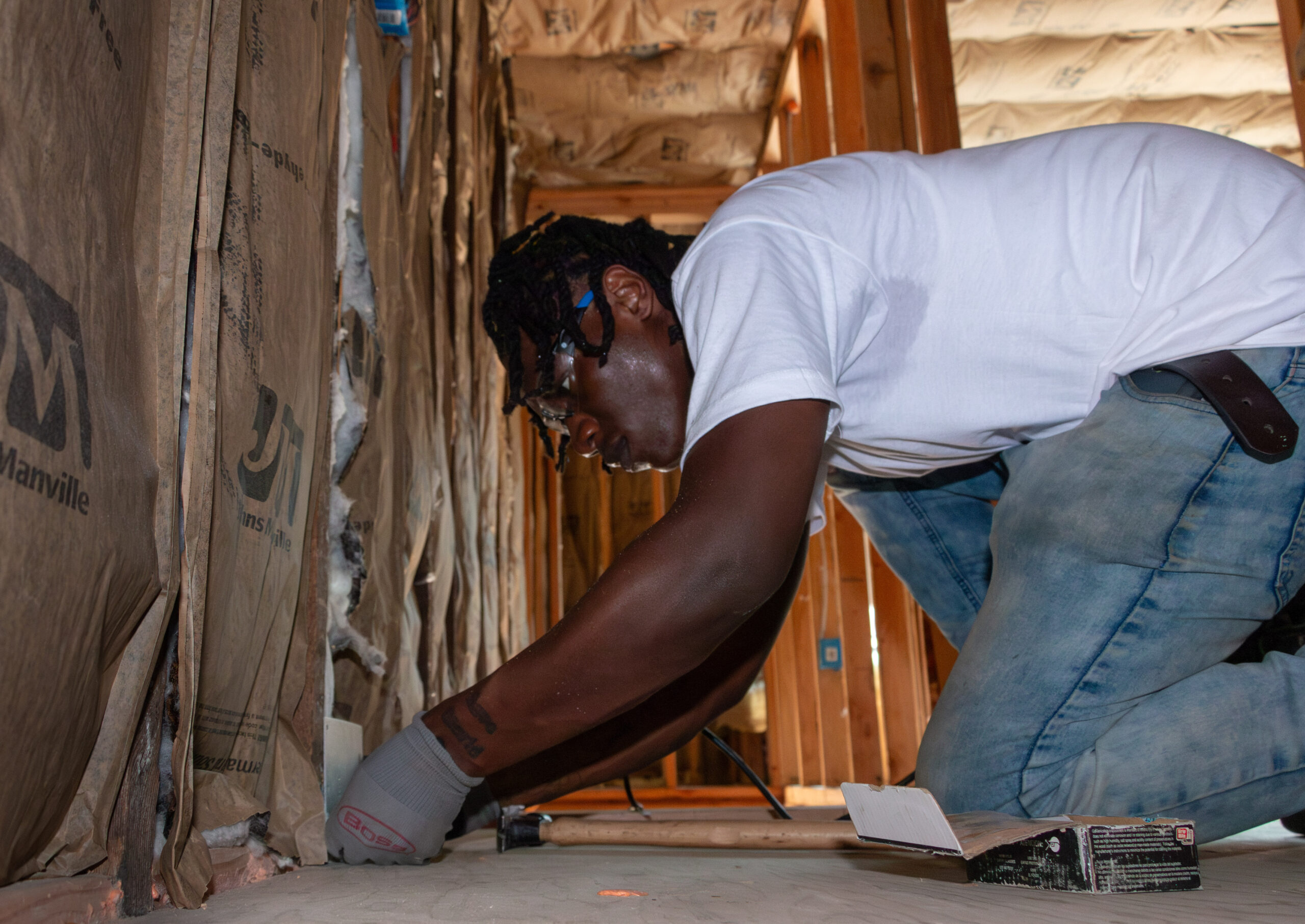 A person wearing a white T-shirt and jeans is kneeling, installing insulation in a wooden-framed room. They appear focused and diligent. Tools are nearby.