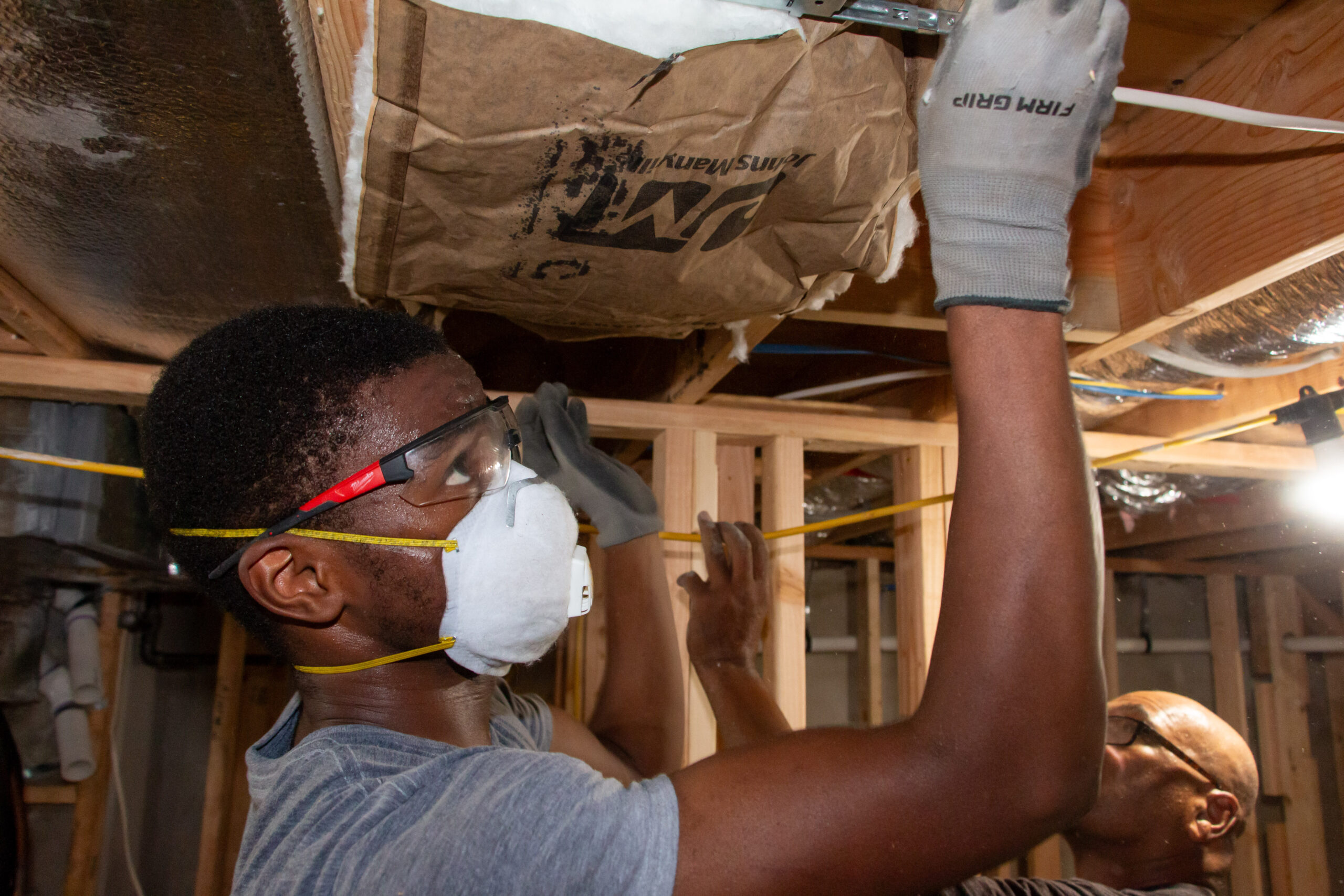 A person wearing safety goggles, a mask, and gloves installs insulation in the ceiling of a room under construction. Another worker is visible in the background.