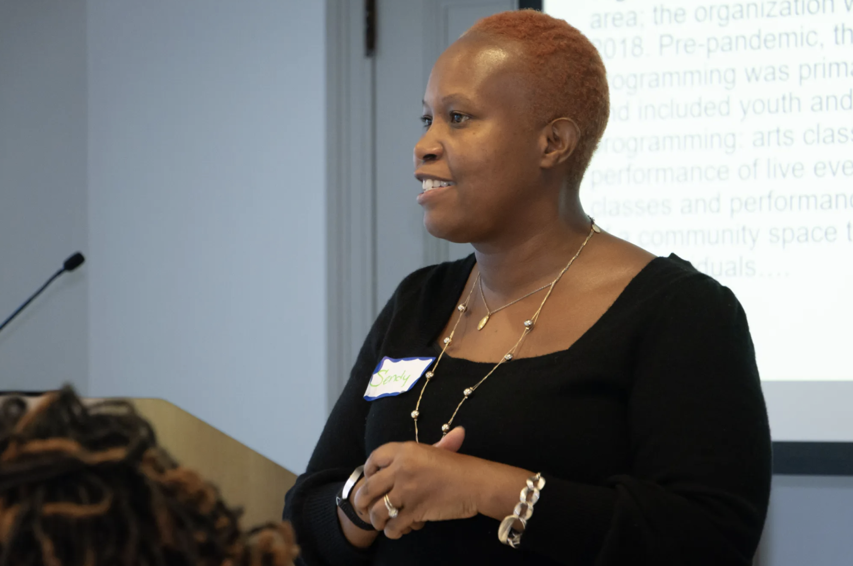A woman with short hair smiles while speaking in a room with a projector in the background. They wear a black top and a name tag, conveying a professional and engaging tone.