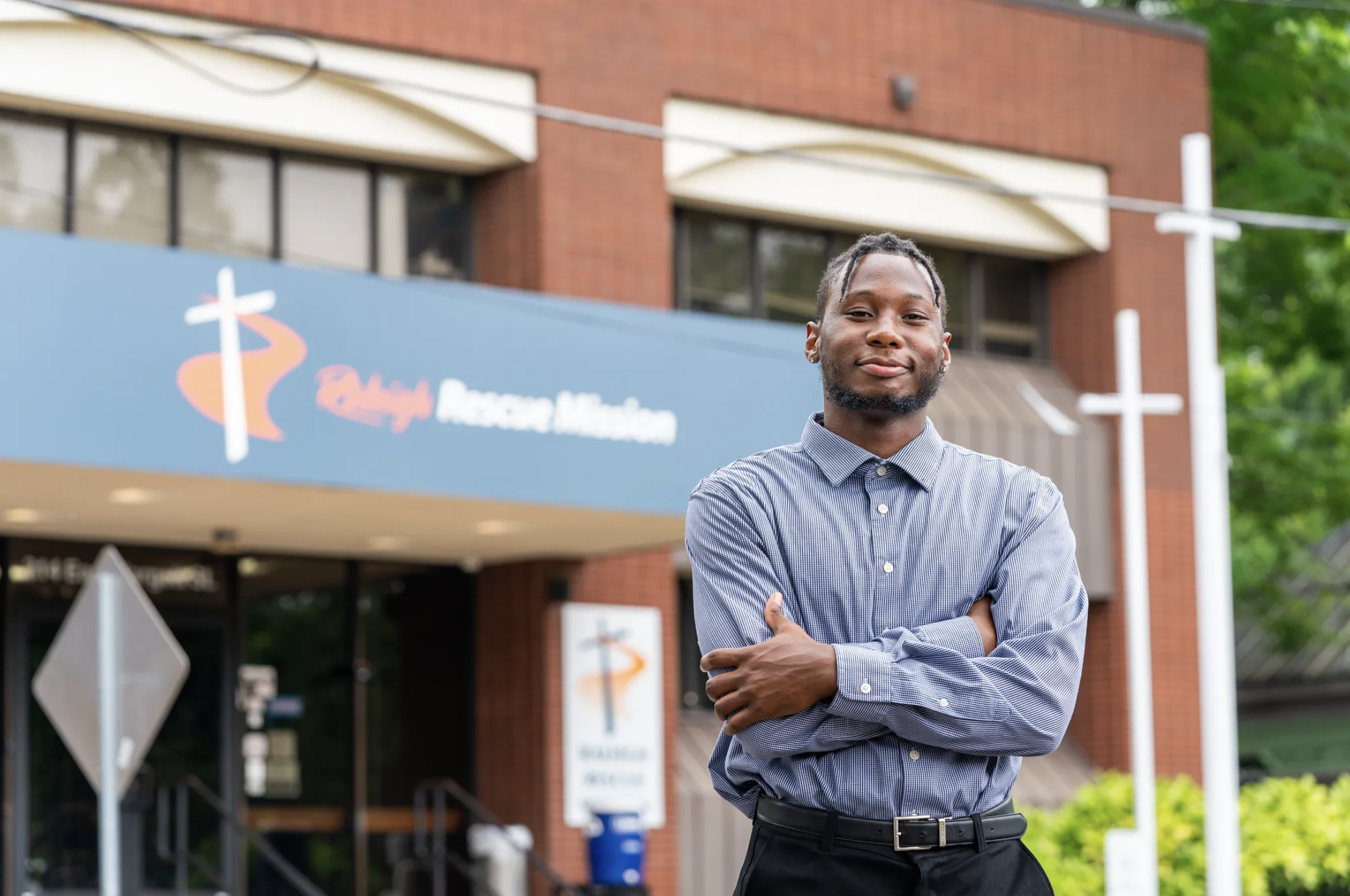 A man in a striped shirt stands confidently with arms crossed in front of a building with a Raleigh Rescue Mission sign. The mood is positive and empowering.