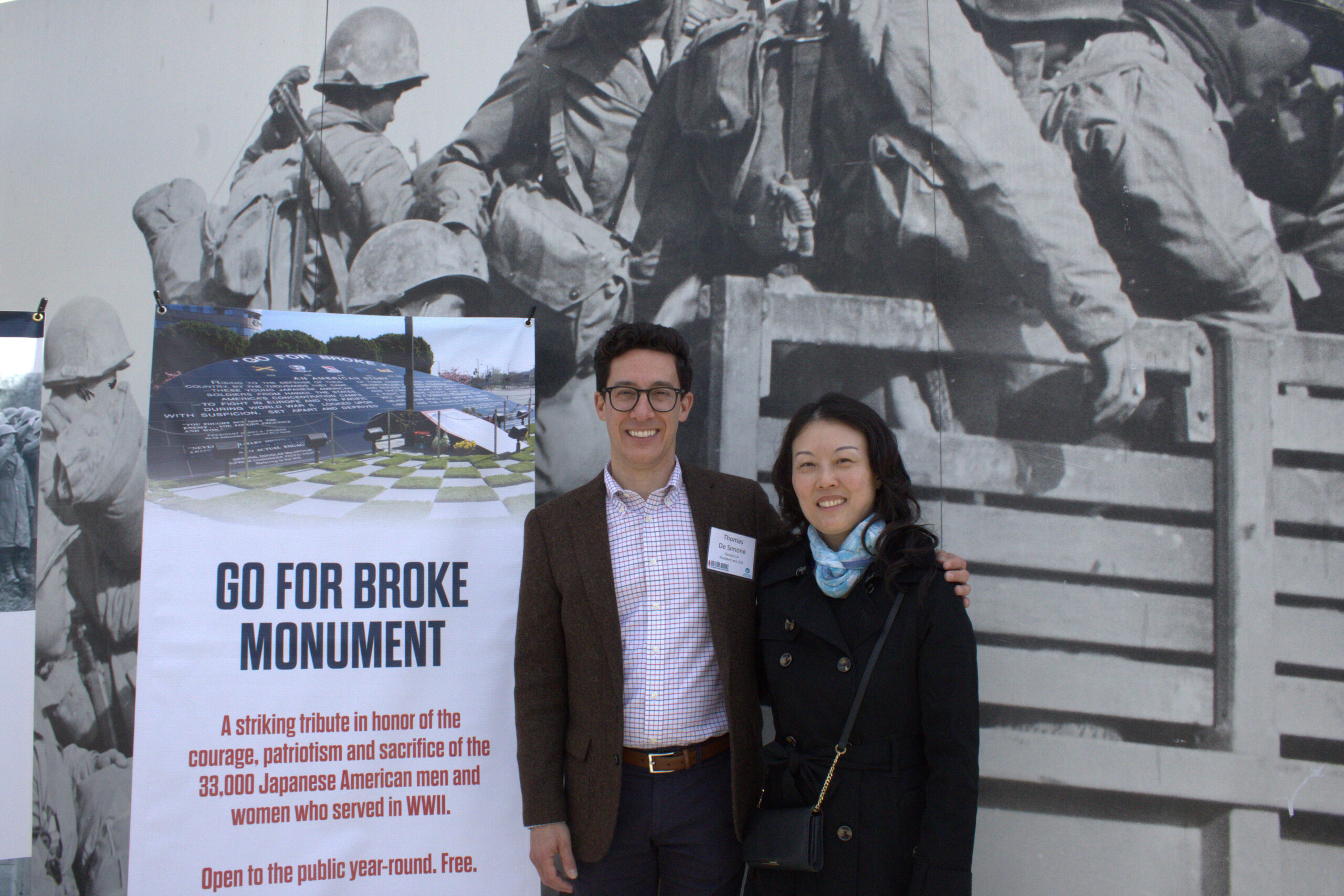 A man and woman smile for a photo, standing next to a large sign for the "Go For Broke Monument." The sign honors the 33,000 Japanese American men and women who served in WWII, and behind them is a black-and-white mural of soldiers.