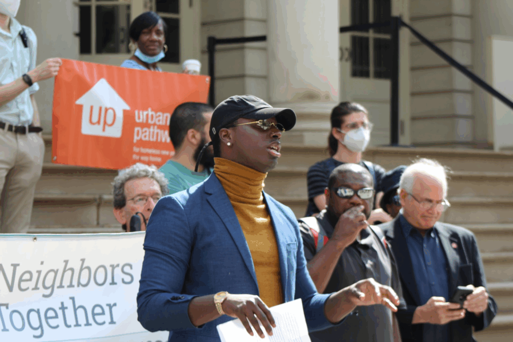 A man in a blue blazer and yellow turtleneck speaks at a press conference on the steps of a building. Behind him, supporters stand in solidarity, holding signs for "Urban Pathways" and "Neighbors Together." conference. Behind him, supporters stand on the steps of a building, holding signs for "Urban Pathways" and "Neighbors Together."