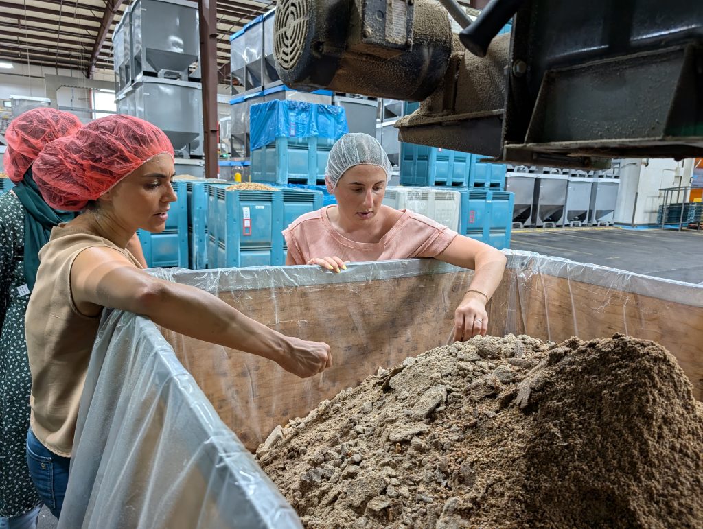 Two women with hairnets touch crushed walnut shells at a walnut processing facility.