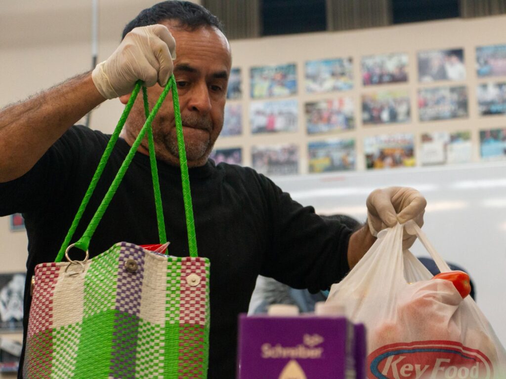 A man wearing disposable gloves handles groceries at a food bank, holding a colorful woven tote and a plastic grocery bag filled with food.